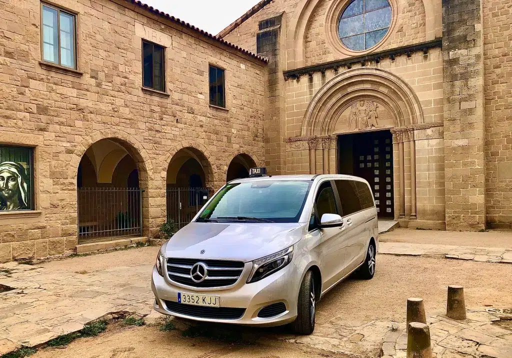 Taxi monovolumen Mercedes estacionado frente a un monasterio románico durante un trayecto de largo recorrido desde el Vallès Oriental
