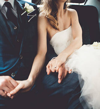 Pareja de novios cogidos de la mano en el asiento trasero de un taxi el día de su boda con el ramo de flores blancas